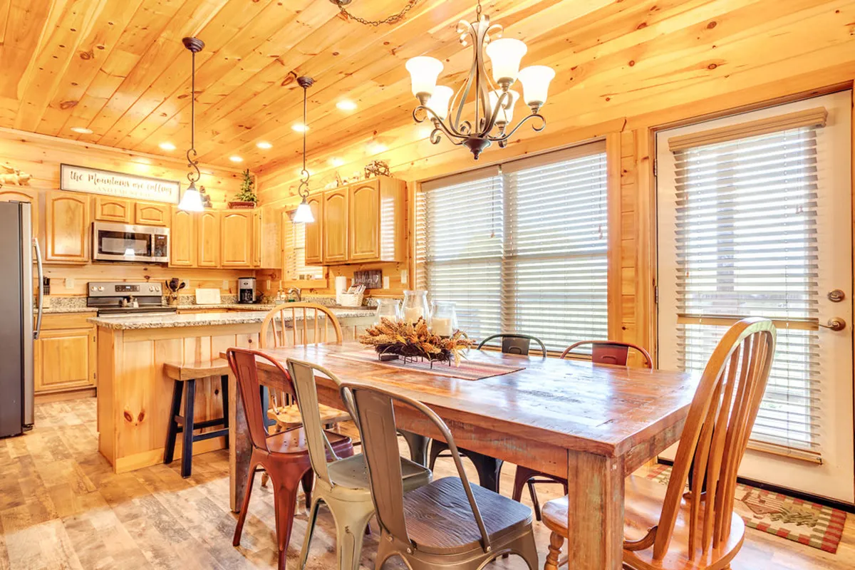 A large wooden dining table set under a chandelier, surrounded by kitchen cabinetry and windows with blinds. This area is welcoming and well-suited to family meals, celebrations, and group entertaining.