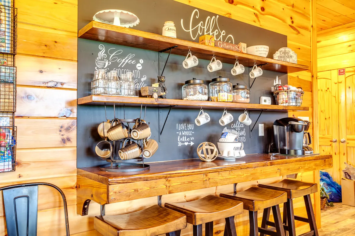 The cozy coffee bar is depicted with wooden shelves holding jars and mugs, and stools inviting guests to linger. The scene emphasizes the ritual of brewing coffee and casual morning conversations.