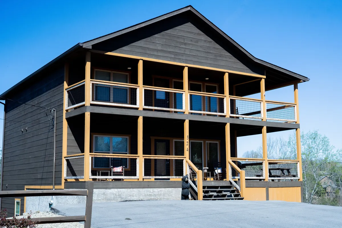 A wide shot of the property's exterior showcases the spacious front porch, balcony, and beautiful landscaping. The wooden structure exudes a welcoming, resort-style vibe, inviting guests to relax outdoors and enjoy the surrounding scenery. The porch and balcony are perfect for family gatherings or quiet mornings with coffee.