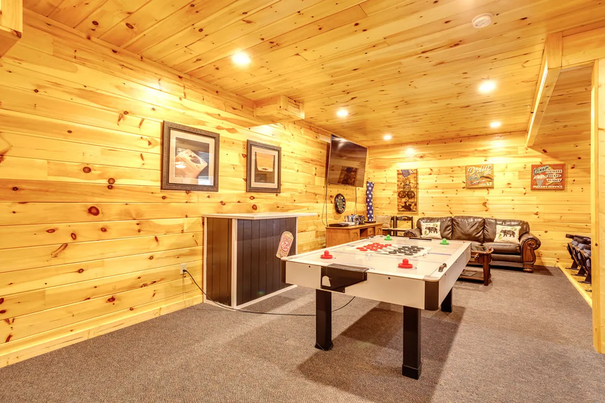 A vibrant game room featuring an air hockey table as the centerpiece, complemented by a comfortable leather couch, wood paneling, wall art, and a television. The room exudes a playful yet relaxed atmosphere, ideal for gatherings and friendly competition.