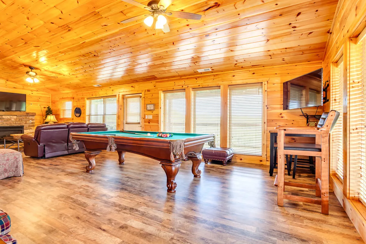 A spacious game room featuring a pool table, comfortable seating, and large windows that let in abundant natural light. The wooden ceiling and decor add a rustic, lodge-like ambiance, emphasizing both leisure and social fun.