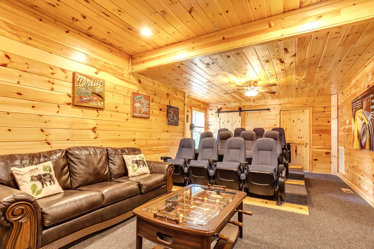 A cozy home theater setup with plush cinema seating, a leather sofa, wooden paneling, and a game table in the foreground. The space is inviting, perfect for immersive movie nights or friendly tabletop competitions.