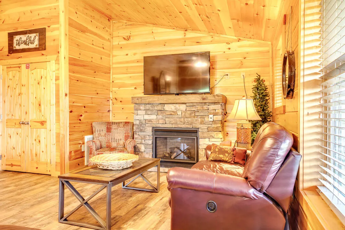 A warmly lit main living area featuring a stone fireplace, a reclining chair, a coffee table, and a television. The room exudes a cozy, inviting ambiance perfect for relaxing by the fire, watching TV, or gathering for game nights.