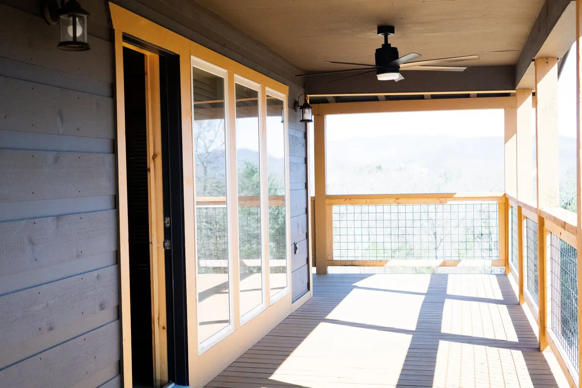 A serene exterior view from the wrap-around deck, featuring large glass doors, wooden railing, and panoramic mountain vistas. The mood is calm and inviting, perfect for enjoying morning coffee or sunsets.