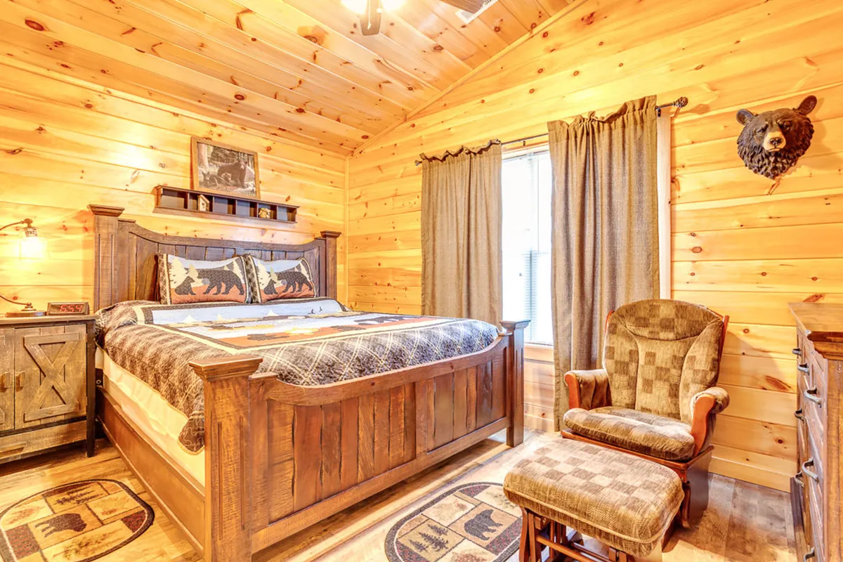 A king suite featuring a solid wooden bed with bear-themed decor, cozy plaid bedding, rustic wooden furniture, and natural wood walls. Gentle sunlight streams in through a nearby window, casting a warm, inviting glow. The atmosphere is tranquil, perfect for relaxing or enjoying a quiet moment alone.